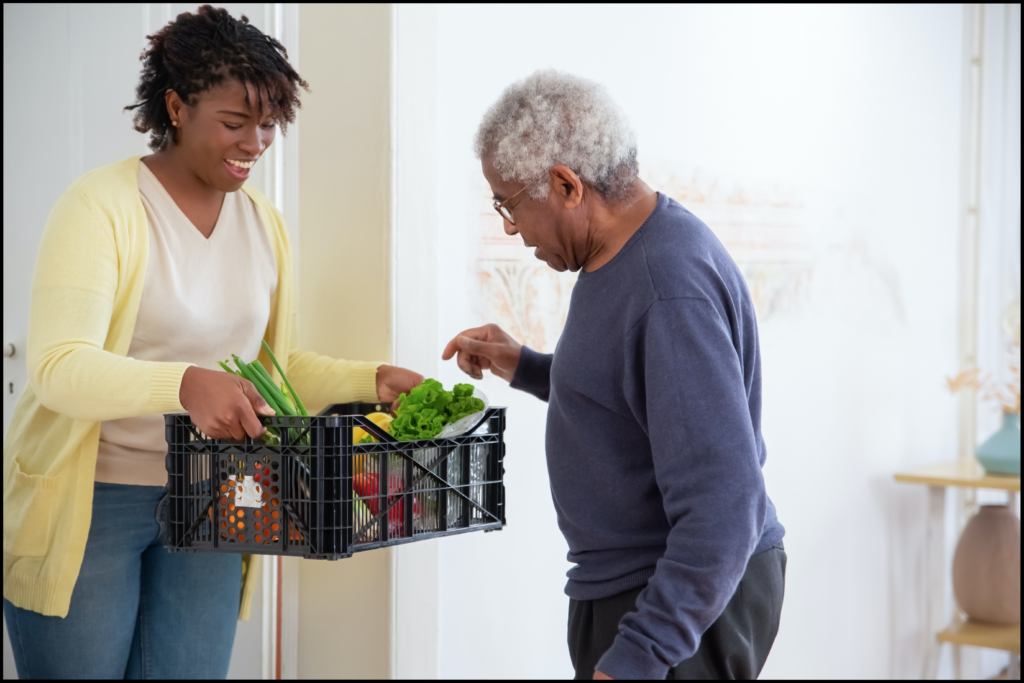 A woman holding a crate of vegetables while an old man picks